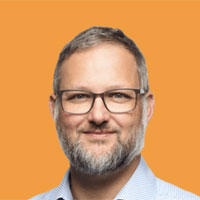 Porträtfoto eines Mannes mit Brille vor orangem Hintergrund, Blick in die Kamera, professionell beleuchtet.