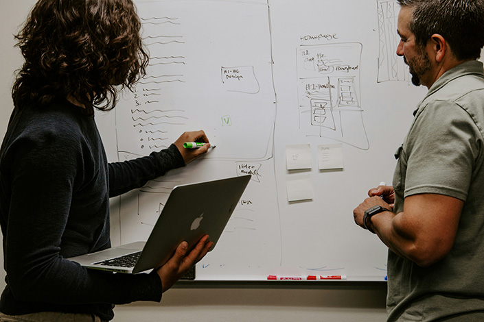 Zwei Männer diskutieren an einem Whiteboard mit Skizzen und Notizen. Einer hält einen Laptop und einen Marker.