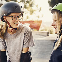 Zwei Mädchen mit Schutzhelmen und Schutzausrüstung beim Gespräch in einer Skatepark-Umgebung, seitliche Ansicht, sonnige Lichtstimmung.
