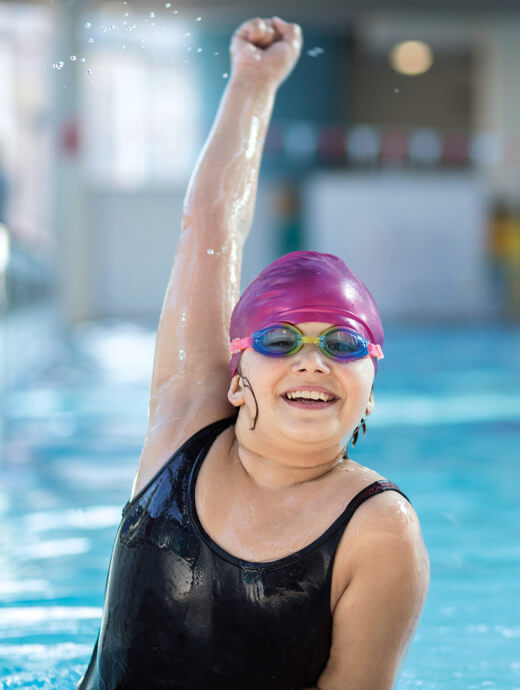 Ein Kind mit Schwimmbrille und Badekappe hebt im Schwimmbecken jubelnd den Arm