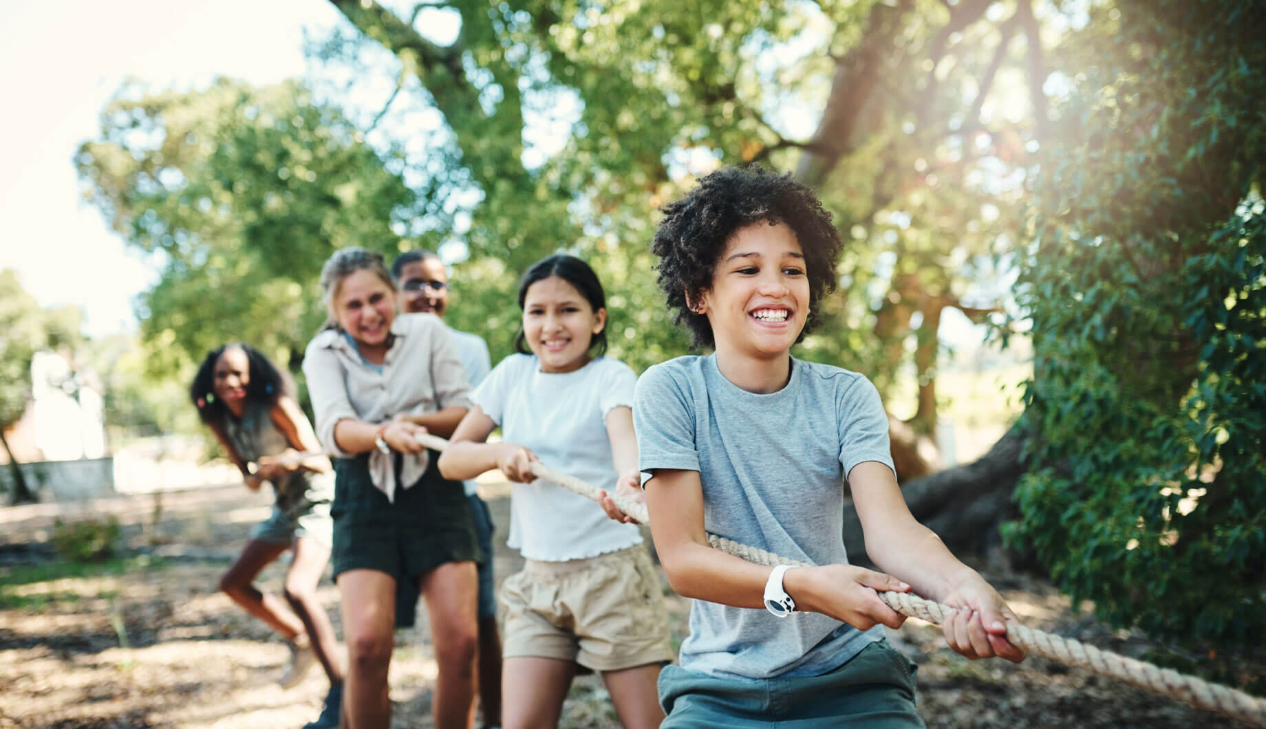 Mehrere Kinder beim Tauziehen auf einem sonnigen Waldspielplatz, im Vordergrund ein lachender Junge in grauem T-Shirt.