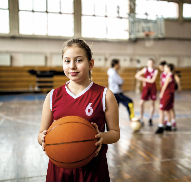 Mädchen im Basketballtrikot mit Ball in einer Turnhalle, im Hintergrund weitere Kinder in Sportkleidung.