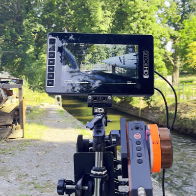 Kameramonitor auf Stativ zeigt Surfszene am Fluss, Aufnahme am Ufer.