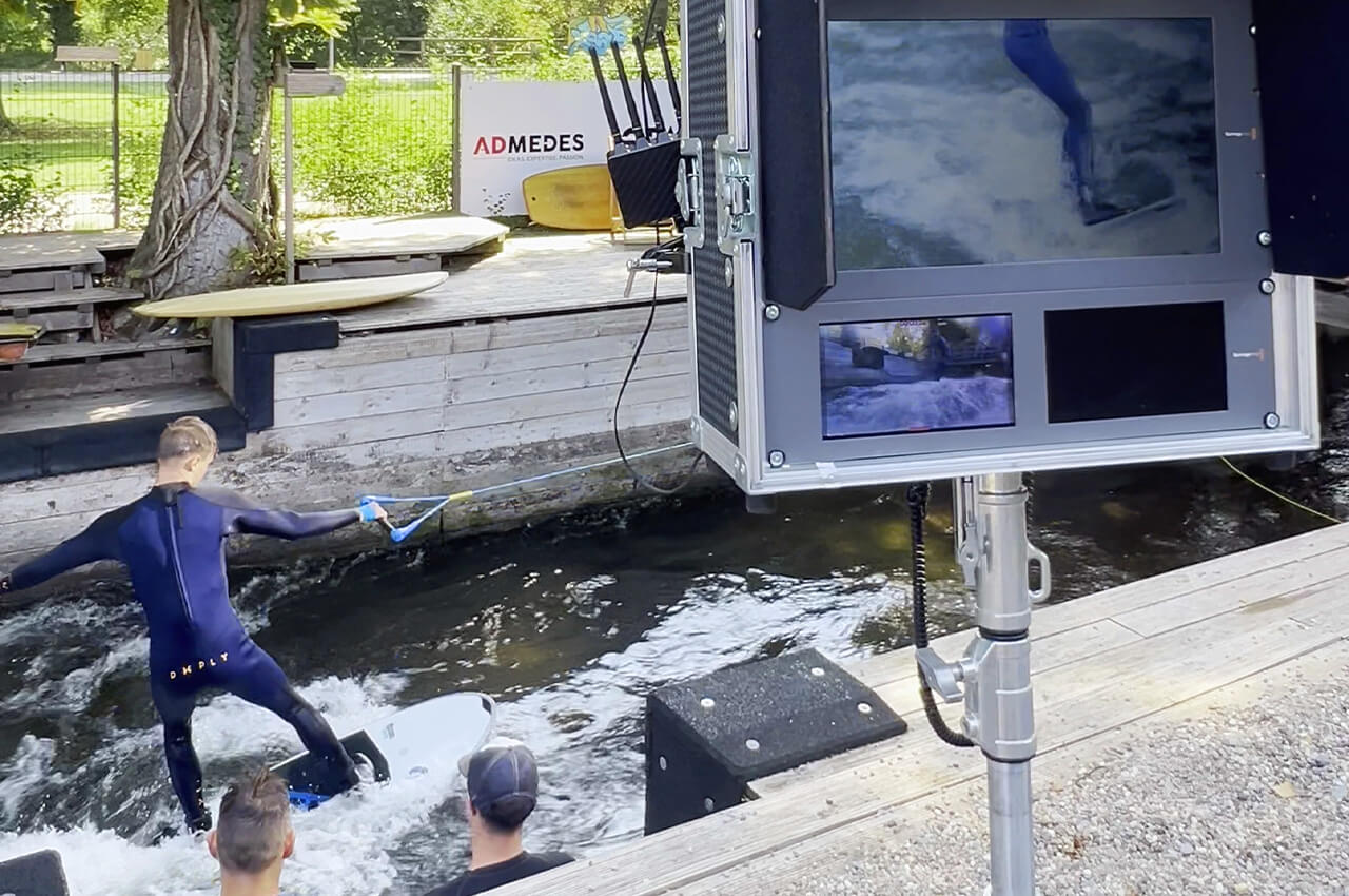 Surfer im Fluss neben Technikaufbau mit Monitoren am Ufer.