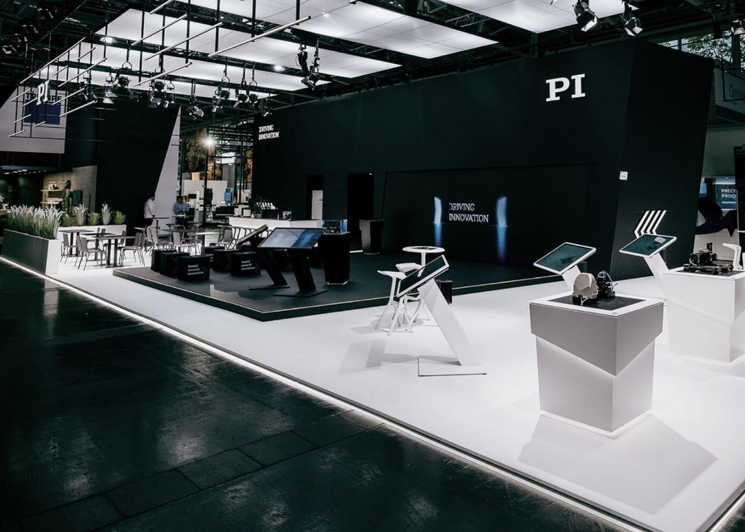 Großer schwarzer Hightech-Messestand von PI mit beleuchtetem Claim „Driving Innovation“, interaktiven Touchscreens, Präsentationsinseln und klarer Zonierung in Weiß und Schwarz.