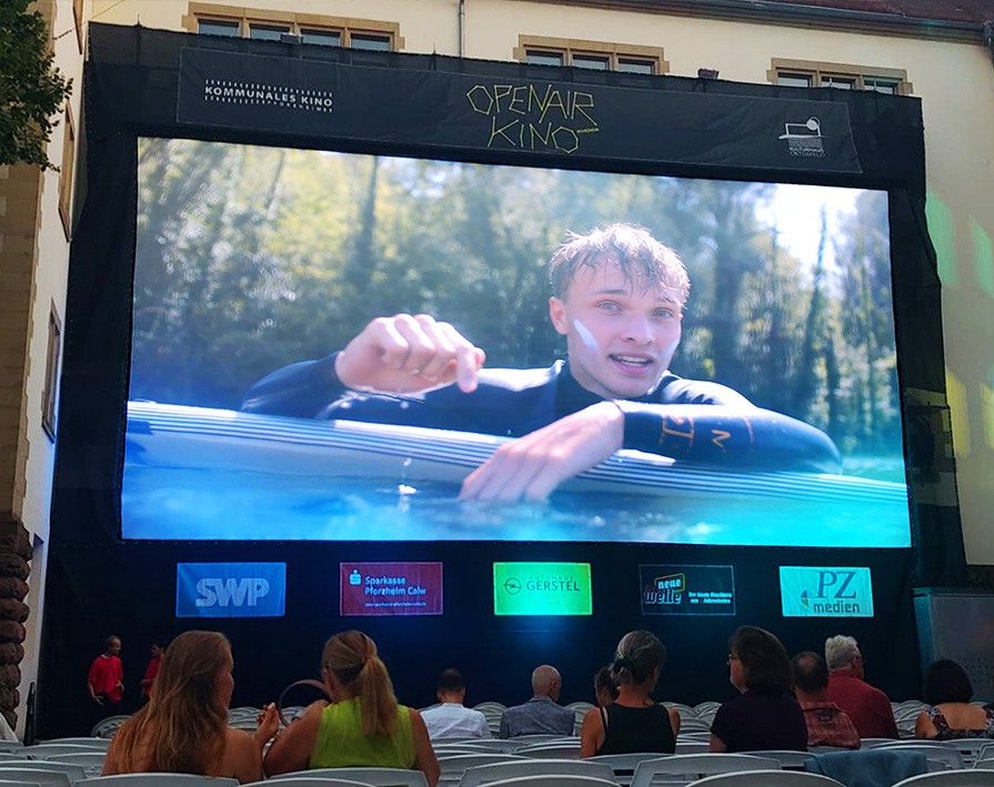 Große Leinwand im Open-Air-Kino zeigt Video von Surfer im Wasser, Publikum sitzt davor.